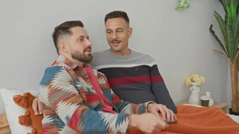 Two men sit on bed under an orange blanket, one points remote at tv while t.. Stock Footage 339217430