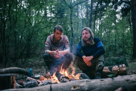Two men sitting around campfire in spring forest. Stock Photos