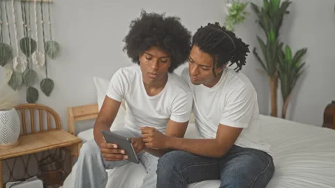 Two men sitting on bed together holding a tablet, one pointing at the scree.. Stock Footage 347694405