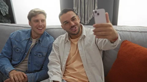 Two men sitting on a couch in a cozy living room laughing and taking a self.. Vídeos de archivo 296030644