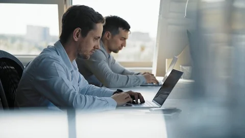 Two men is sitting in front of laptops. One of them uses mobile phone 스톡 동영상 117535758