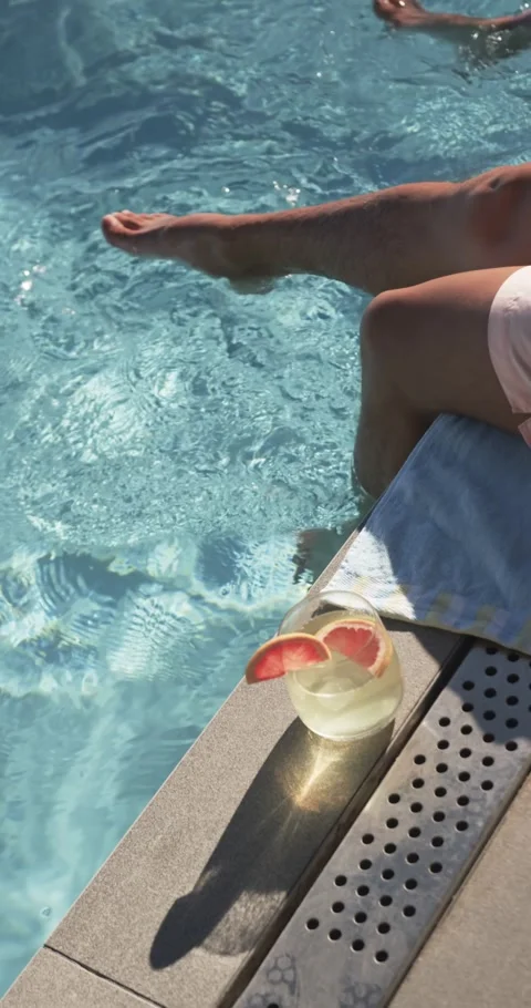 Two men sitting on pool edge, one dipping foot creating ripples, glass on towel Stock Footage 345322475