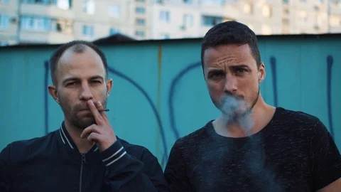 smoking two cigarettes Two men smoke cigarettes in the ghetto and look into the camera