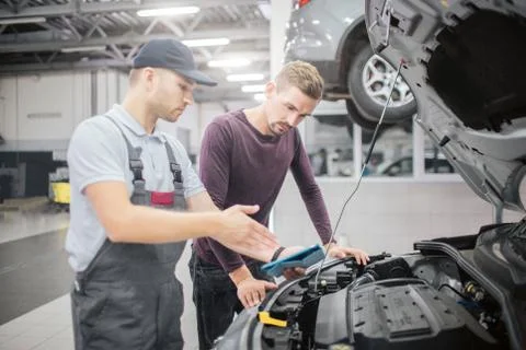 Two men stand at opened front part of car body. Worker point on it. They have Stock Photos
