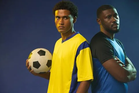 Two men standing back-to-back in studio holding soccer ball, wearing yellow Stock Photos
