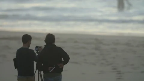 Two men standing at beach and recording ocean waves with camera on tripod Stock Footage 158632684