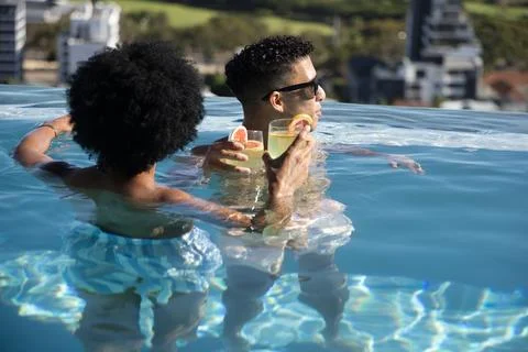 Two men standing in rooftop infinity pool with bare torsos holding stemless Stock Photos
