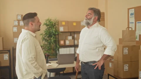 Two men standing by stacked parcels and a laptop, one arms crossed and the .. Stock Footage 339214069