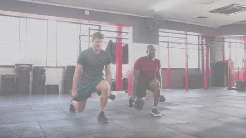Two men starting side-by-side, doing in-sync dumbbell walking lunges across gym Stock Footage 334203814