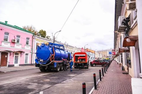 Two men, utility worker and other, connect large blue sewage truck with hose to Stock Photos