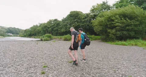 Two men walking along rocky riverbank, enjoying nature and outdoor exercise Stock Footage 346028501