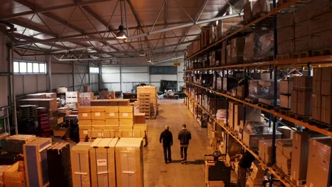 Two men walking through a storage warehous area with stacked boxes. 스톡 동영상 166981430