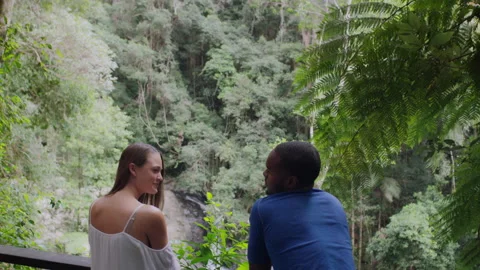 Two millennial friends leaning on railing smiling in front of small waterfall in Video stock 199463599