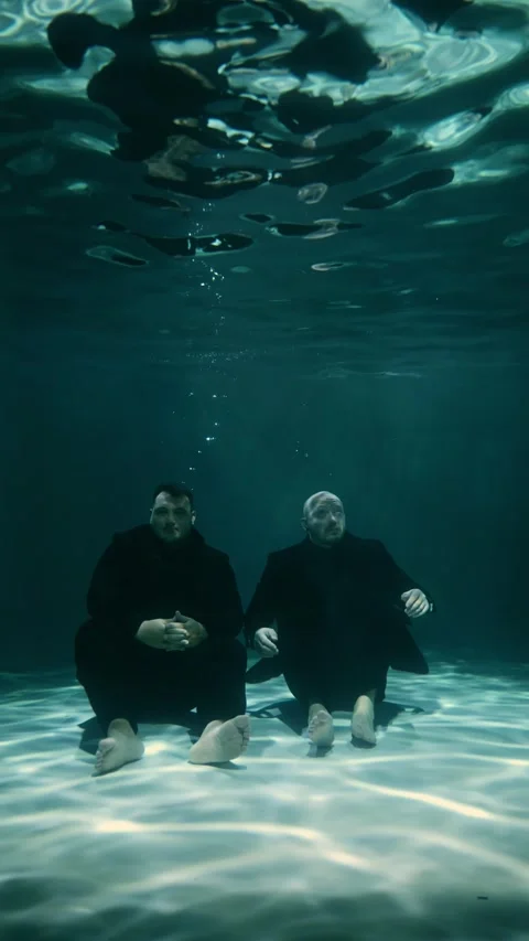 Two millennial men in black suits are sitting at the bottom of a swimming pool Stock Footage 315559293