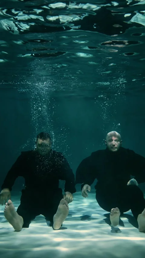Two millennial men in black suits sink to the bottom of the pool and sit down Stock Footage 315560097
