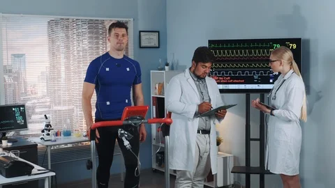 Two mixed race doctors discussing EKG data of athlete doing treadmill test in Stock Footage 114081351