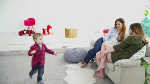 Two mothers sitting on the couch talking while their children running around the Stock Footage 75016495