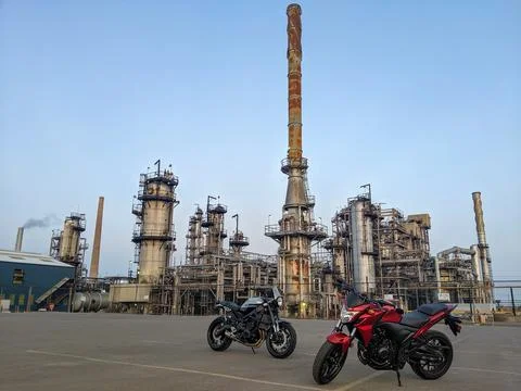 Two Motorcycles Parked in Front of Rusty Industrial Refinery Chimney at Dusk Foto stock