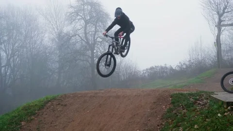Two mountain bikers jump over a  jump in a bike park on a foggy morning 库存影片 234204200