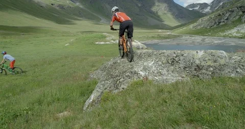 Two mountain bikers ride over a rock face in high mountains and lakes. Video stock 78030457