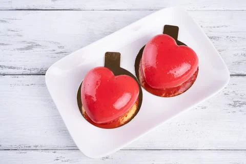 Two mousse cakes in the form of a red heart on a white plate on white wooden Stock Photos
