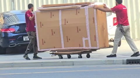 Two movers in red shirts carefully push a large wooden crate on a dolly 스톡 동영상 311885529