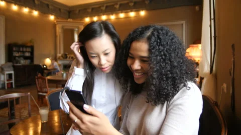 Two Multi Ethnic Girls Having Fun Using Smart Phone Technology In Cafe Видео 98589565
