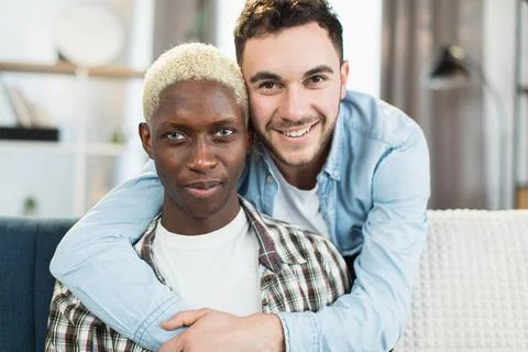 Two multicultural gays embracing and smiling on couch Stock Photos