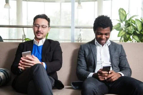 Two multicultural men sit on sofa and hold mobile phones Photos