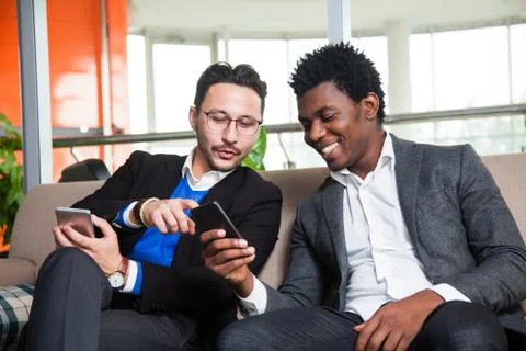 Two multicultural men sit on sofa, smile and hold mobile phones Stock Photos