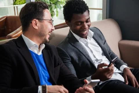 Two multicultural men sit on sofa, smile and hold mobile phones Stockfoto's