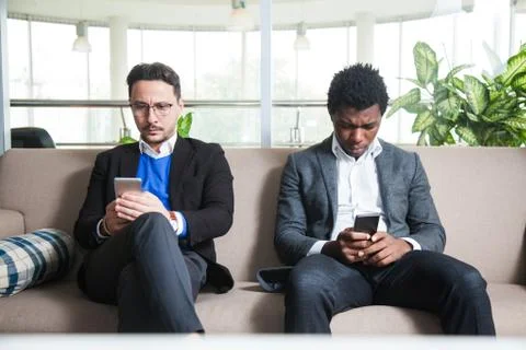 Two multicultural men sit on sofa and hold mobile phones 库存照片