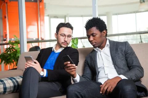 Two multicultural men sit on sofa, smile and hold mobile phones 写真素材