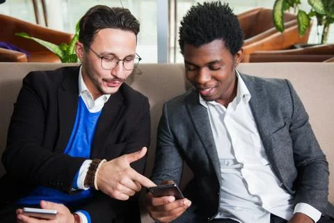 Two multicultural men sit on sofa, smile and hold mobile phones 写真素材