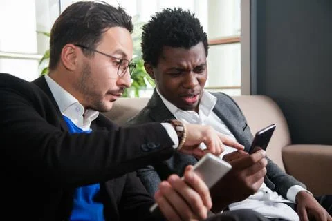 Two multicultural men sit on sofa, smile and hold mobile phones Fotos Stock