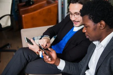 Two multicultural men sit on sofa, smile and hold mobile phones Photos