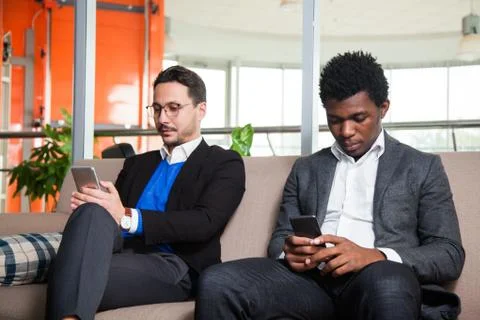 Two multicultural men sit on sofa, smile and hold mobile phones Fotos de archivo