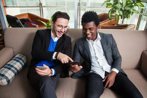 Two multicultural men sit on sofa, smile and hold mobile phones 写真素材