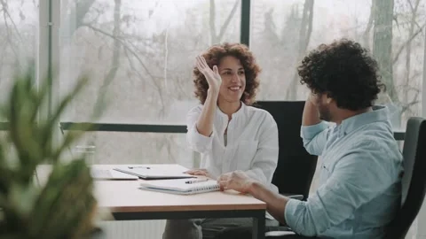 Two multiethnic employees sitting in the office at the desk give each other the Stock Footage 240405796