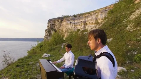 Two musicians Pianist and accordionist playing on the rocks at sunset over sea Stock Footage