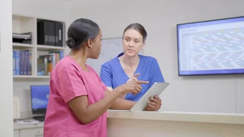 Two nurses at nurses station review patient chart on iPad, camera move right Stock Footage 109869921