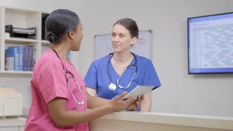 Two nurses w/ stethoscopes at nurses station review chart, camera move right Stock Footage 109869066