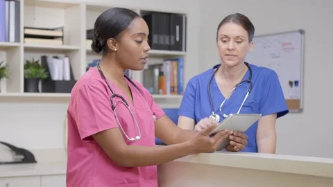 Two nurses w/ stethoscopes at nurses station review chart, camera move left Stock Footage 109869701