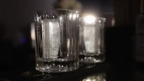 Two Old Fashioned glasses are standing on the bar counter with a cube of ice Stock Footage 240303273