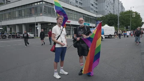 Two older men hold rainbow flags while standing on a street as participants pass Stock Footage 346238623