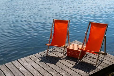 Two orange deck chairs sit on a wooden dock by a calm lake Stock Photos