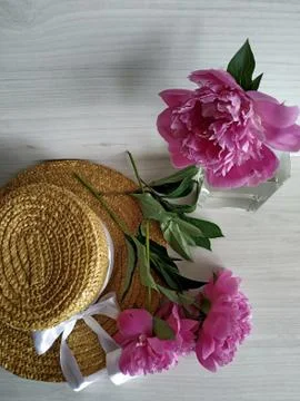 Two peonies on a wicker straw hat and one peony standing in a transparent vase Stock Photos