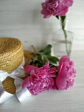 Two peonies on a wicker straw hat and one peony standing in a transparent vase Stock Photos