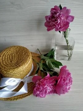 Two peonies on a wicker straw hat and one peony standing in a transparent vase Stock Photos
