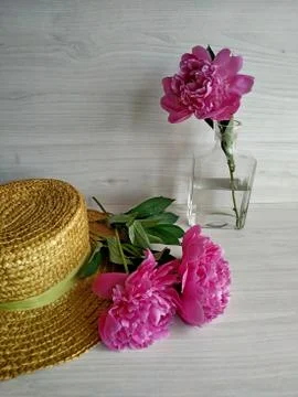 Two peonies on a wicker straw hat and one peony standing in a transparent vase Stock Photos